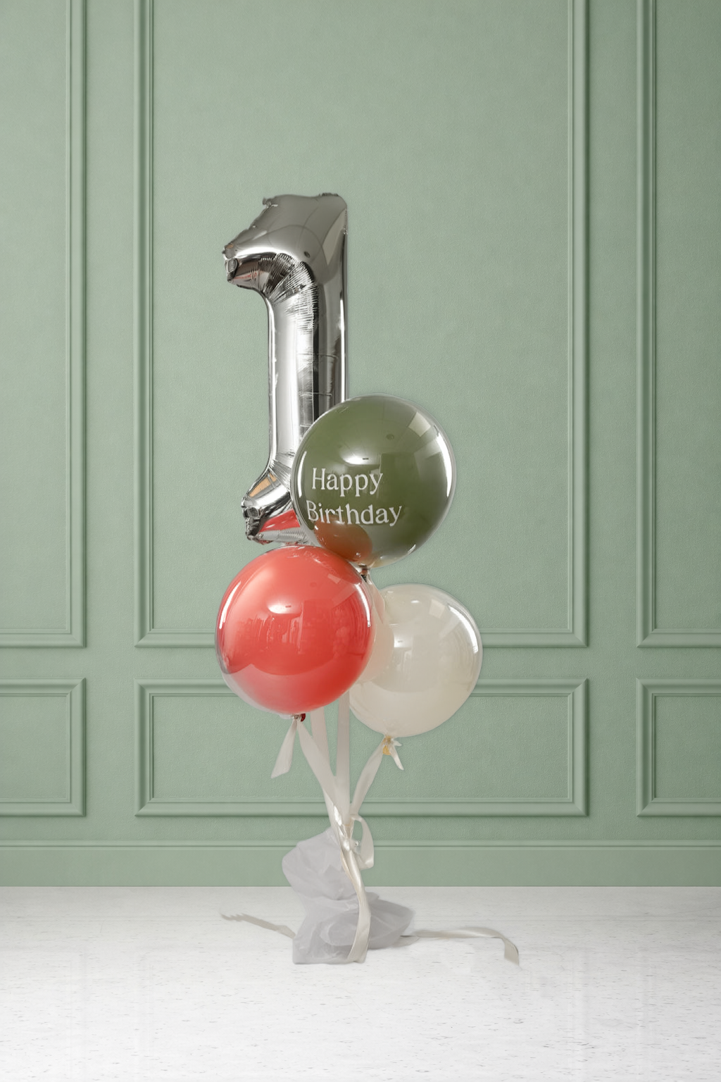 Silver number balloon shaped like '1' above a bunch of three orbz balloons — olive green printed with 'Happy Birthday,' coral, and cream — tied with white ribbon and tissue paper to a weight, set against a sage green paneled wall.