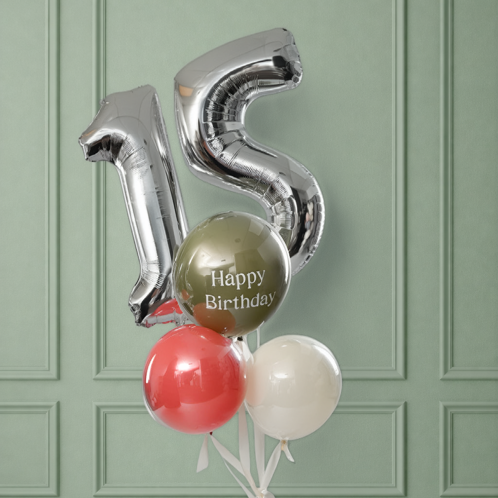Silver number balloons spelling '15' above a bunch of three orbz balloons — olive green printed with 'Happy Birthday,' coral, and cream — tied with white ribbon and tissue paper to a weight, set against a sage green paneled wall.