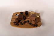 A wooden serving board with chocolate fudge bars, chocolate chips, and four small cups of different colored beverages, photographed against a plain off-white background.