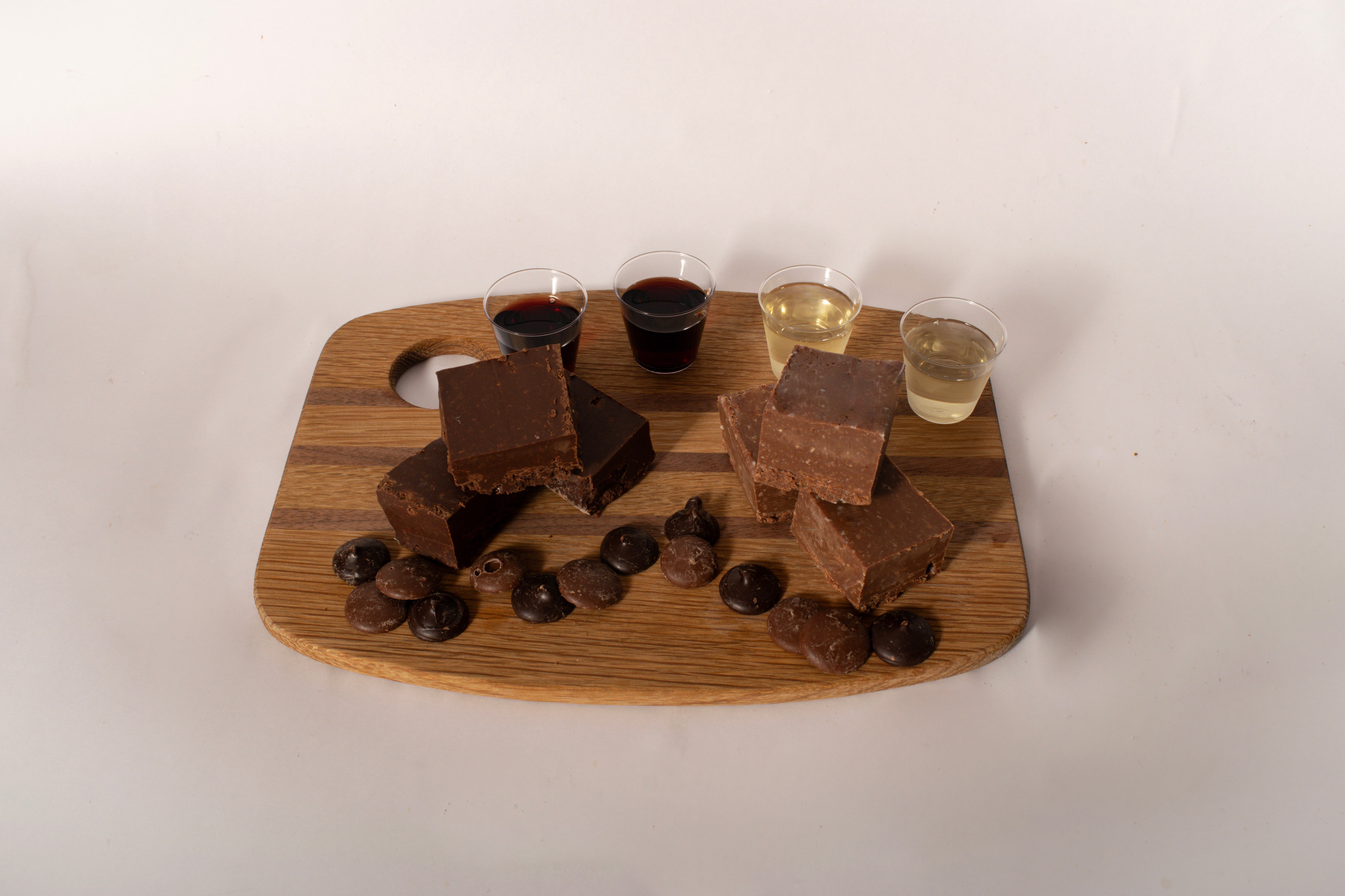 A wooden serving board with chocolate fudge bars, chocolate chips, and four small cups of different colored beverages, photographed against a plain off-white background.