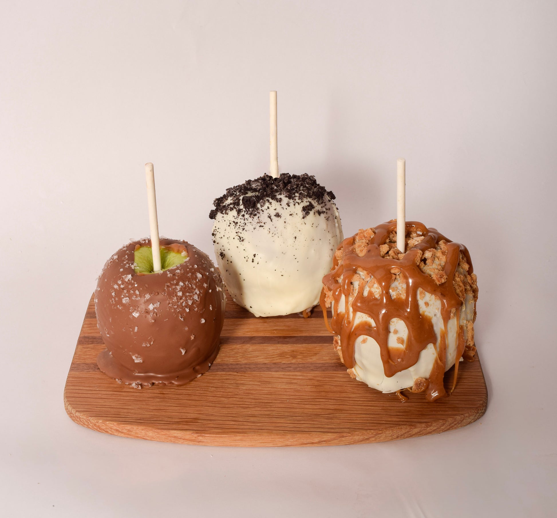 Three chocolate-dipped caramel apples on sticks — milk chocolate with sea salt, white chocolate with crushed cookie topping, and white chocolate with caramel drizzle and streusel crumble — displayed on a wood board.