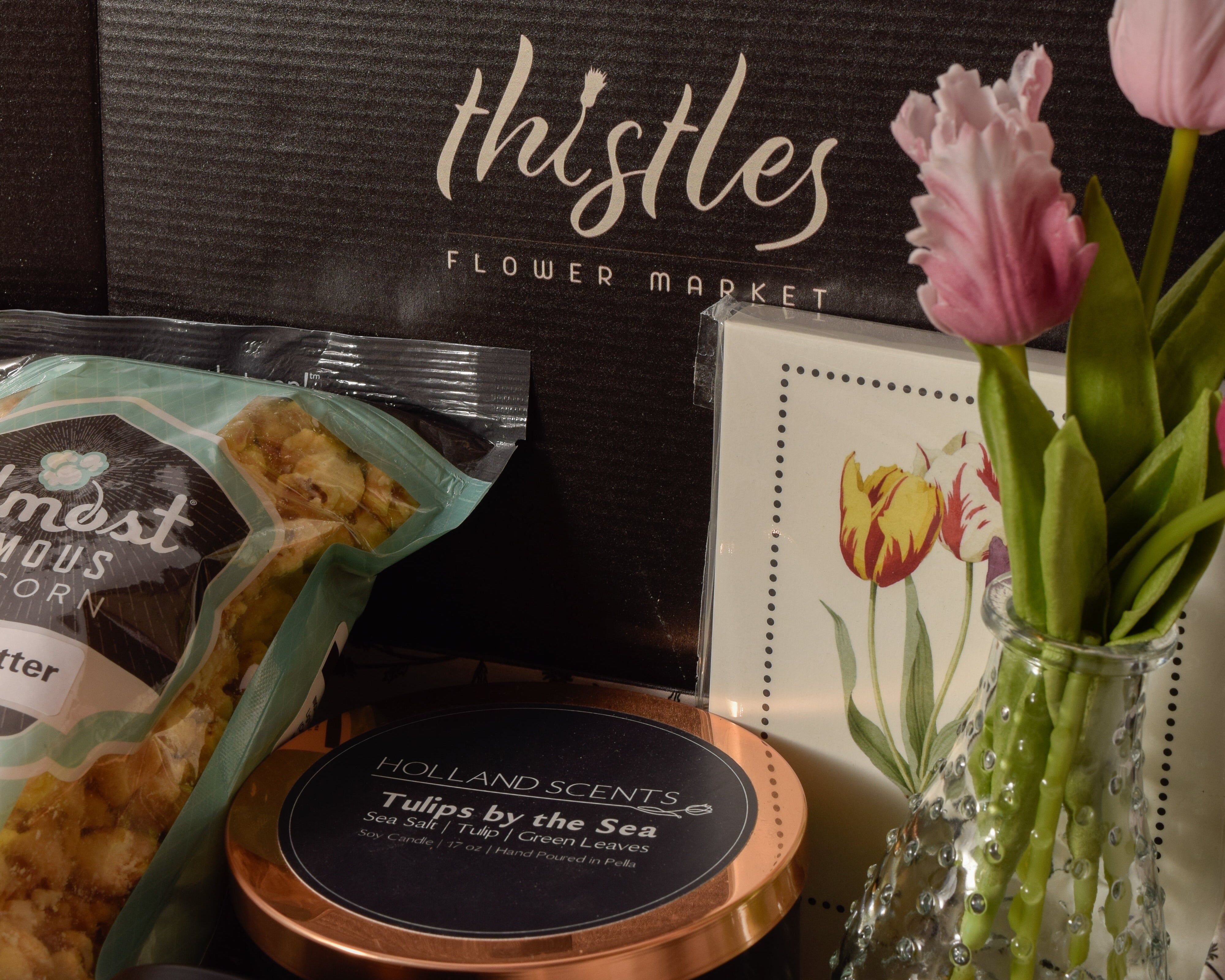 A curated gift box scene with a large black box labeled 'thistles FLOWER MARKET' in the background, alongside a bag of popcorn, a round candle tin, a small jar, and a glass vase holding pink tulips.