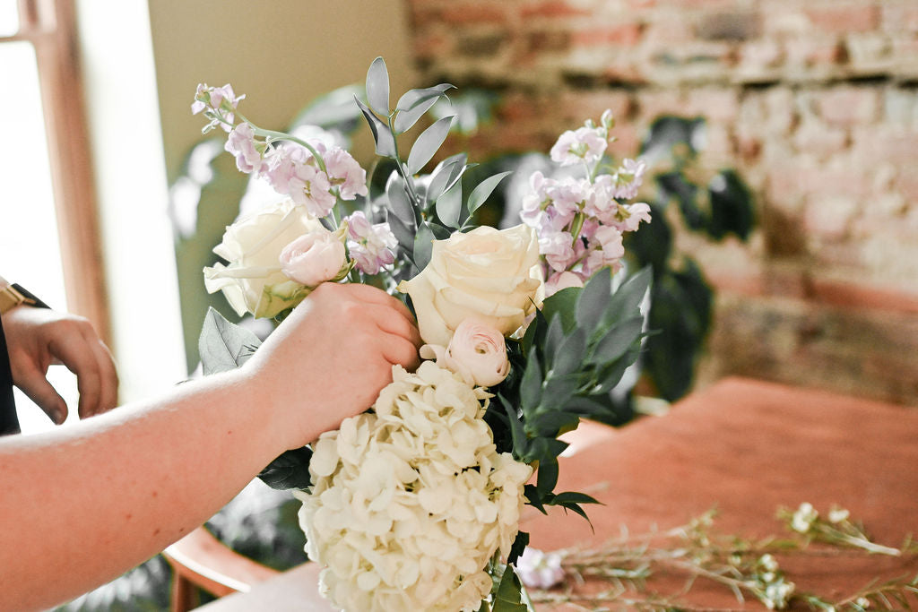 A close-up of a hand arranging a floral bouquet with cream roses, pale pink flowers, green foliage, and a large cream hydrangea, set against a softly blurred indoor background with a brick wall and wooden table.