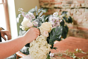 A close-up of a hand arranging a floral bouquet with cream roses, pale pink flowers, green foliage, and a large cream hydrangea, set against a softly blurred indoor background with a brick wall and wooden table.