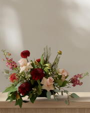Low, wide Thanksgiving floral centerpiece with burgundy dahlias, blush roses, cream lilies, pink stock, and eucalyptus greenery in a low black vase, styled on a wood table.